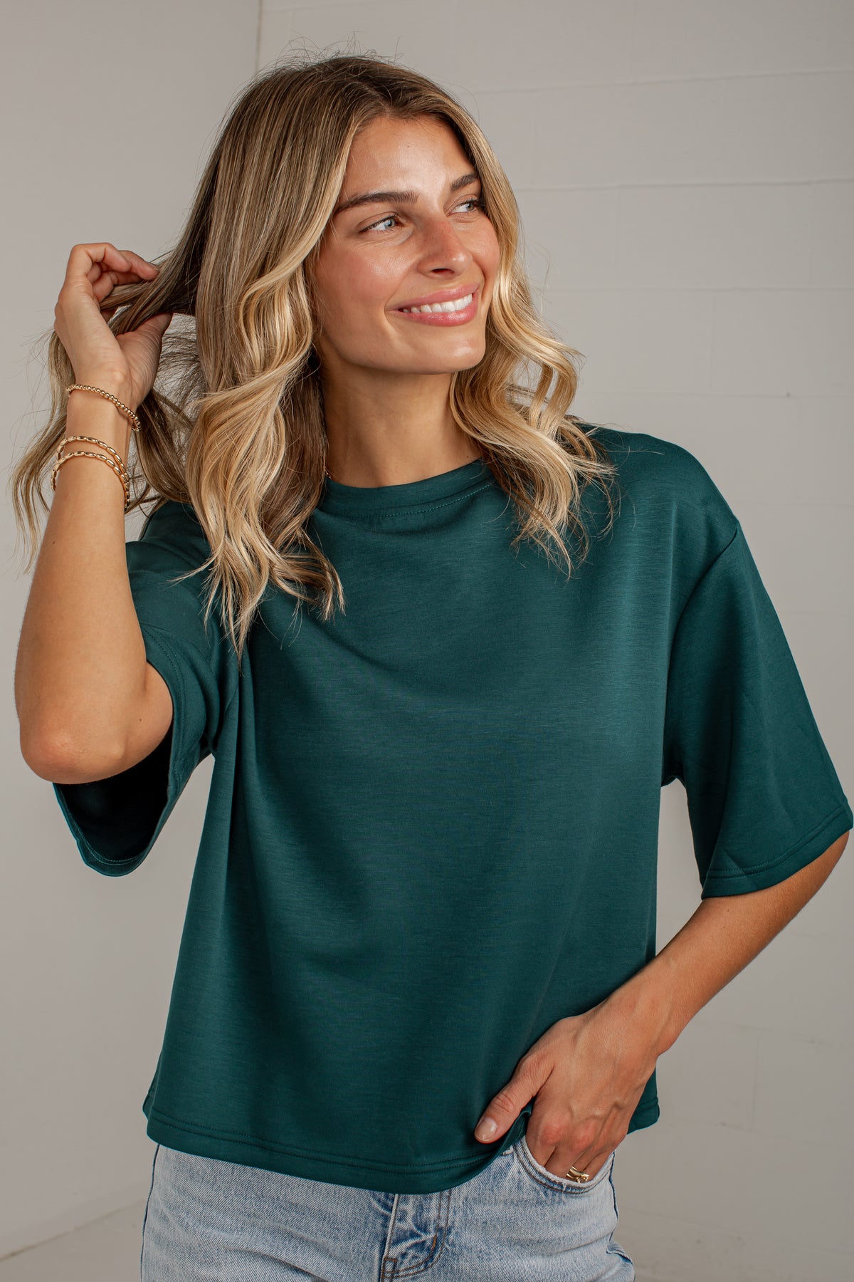 A woman with blonde wavy hair wearing a dark teal green short-sleeve t-shirt and light wash jeans, posing against a light gray paneled wall.