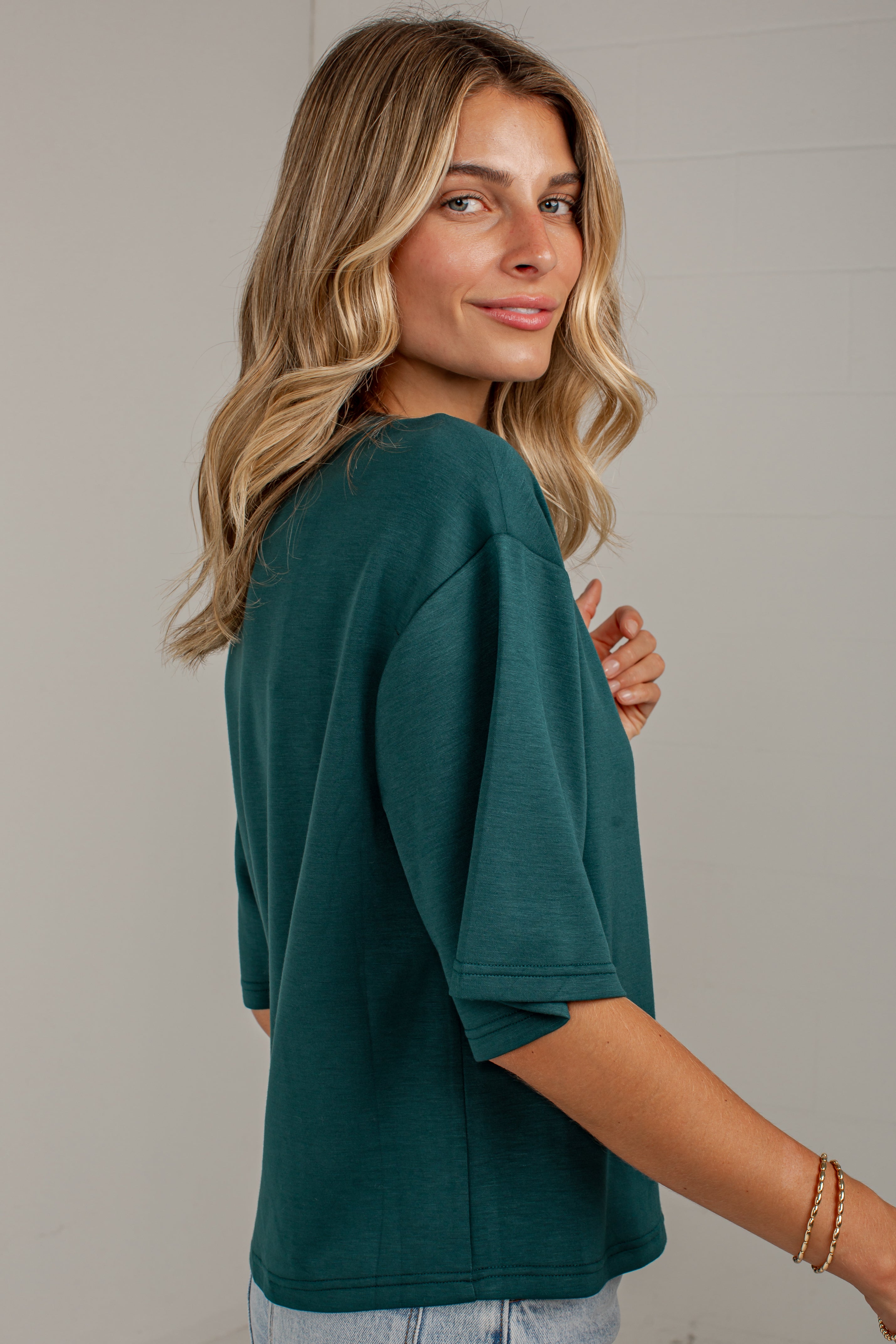 A woman with blonde wavy hair wearing an oversized dark teal green short-sleeve t-shirt, shown from a three-quarter back view against a light gray wall.