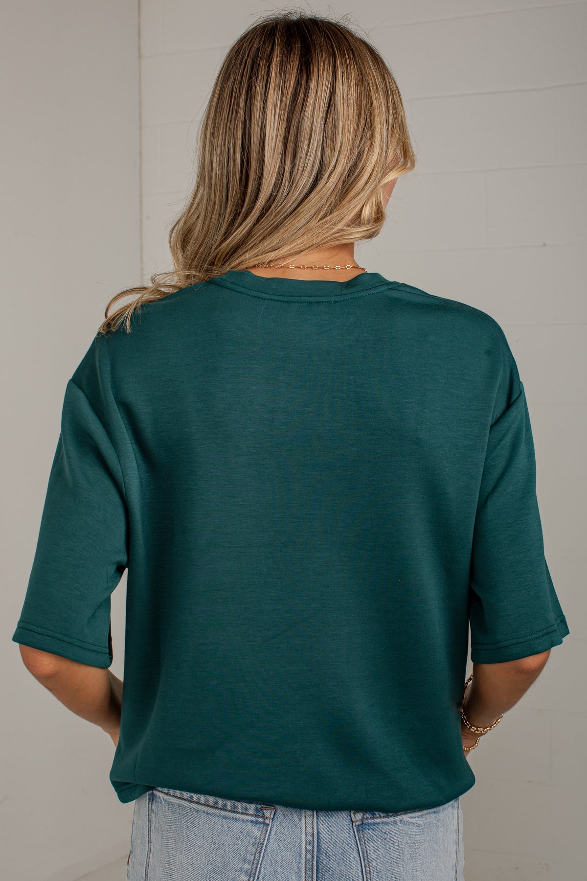 A woman viewed from behind wearing a dark teal green short-sleeve t-shirt and light wash blue jeans, standing against a white paneled wall.