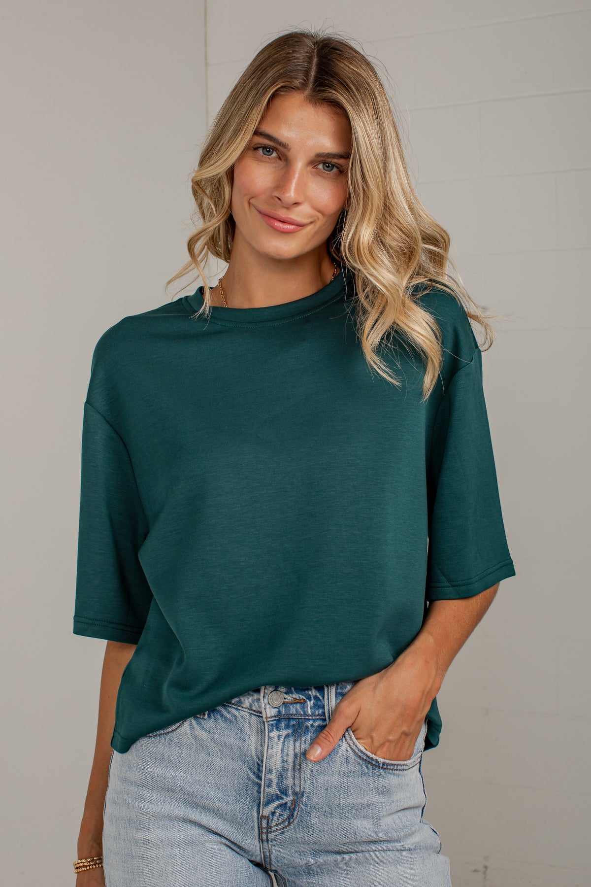 A woman wearing a dark green short-sleeve crewneck t-shirt and light wash blue jeans, standing against a light gray wall.