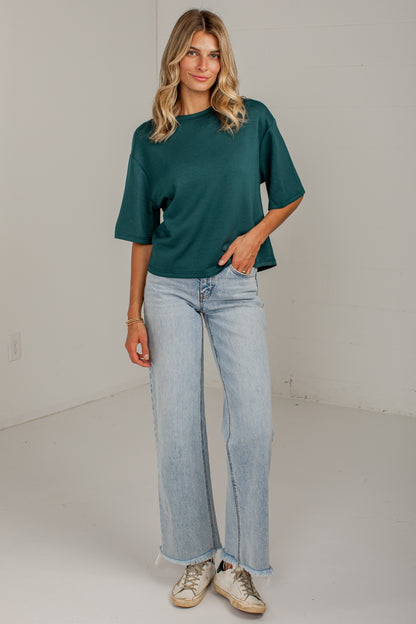 A woman modeling a dark green short-sleeve crewneck t-shirt paired with light-wash wide-leg jeans and beige sneakers, standing against a plain off-white wall.