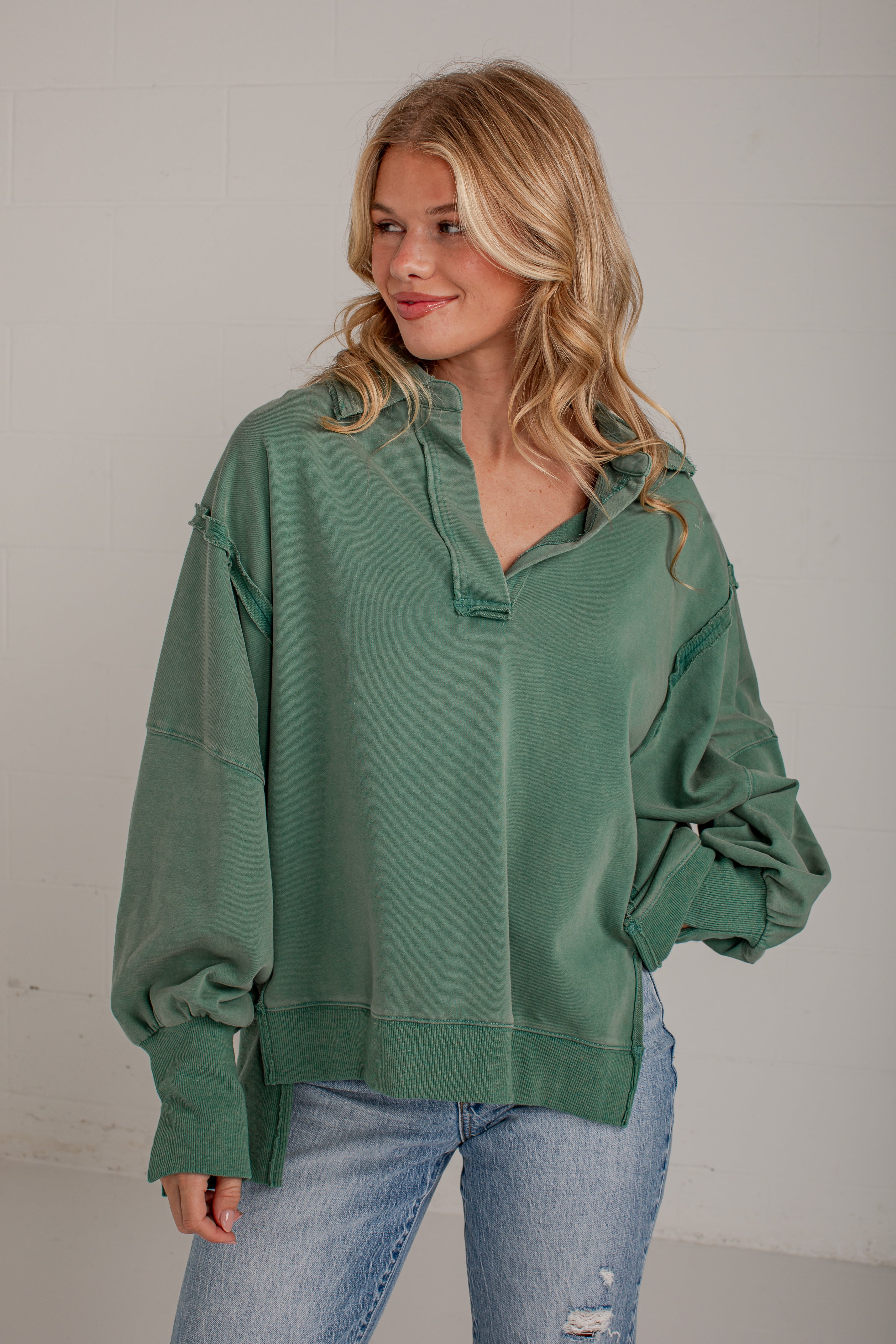 A woman wearing an oversized sage green half-zip hoodie sweatshirt with balloon sleeves and distressed details, paired with light-wash blue jeans, standing against a light gray wall.