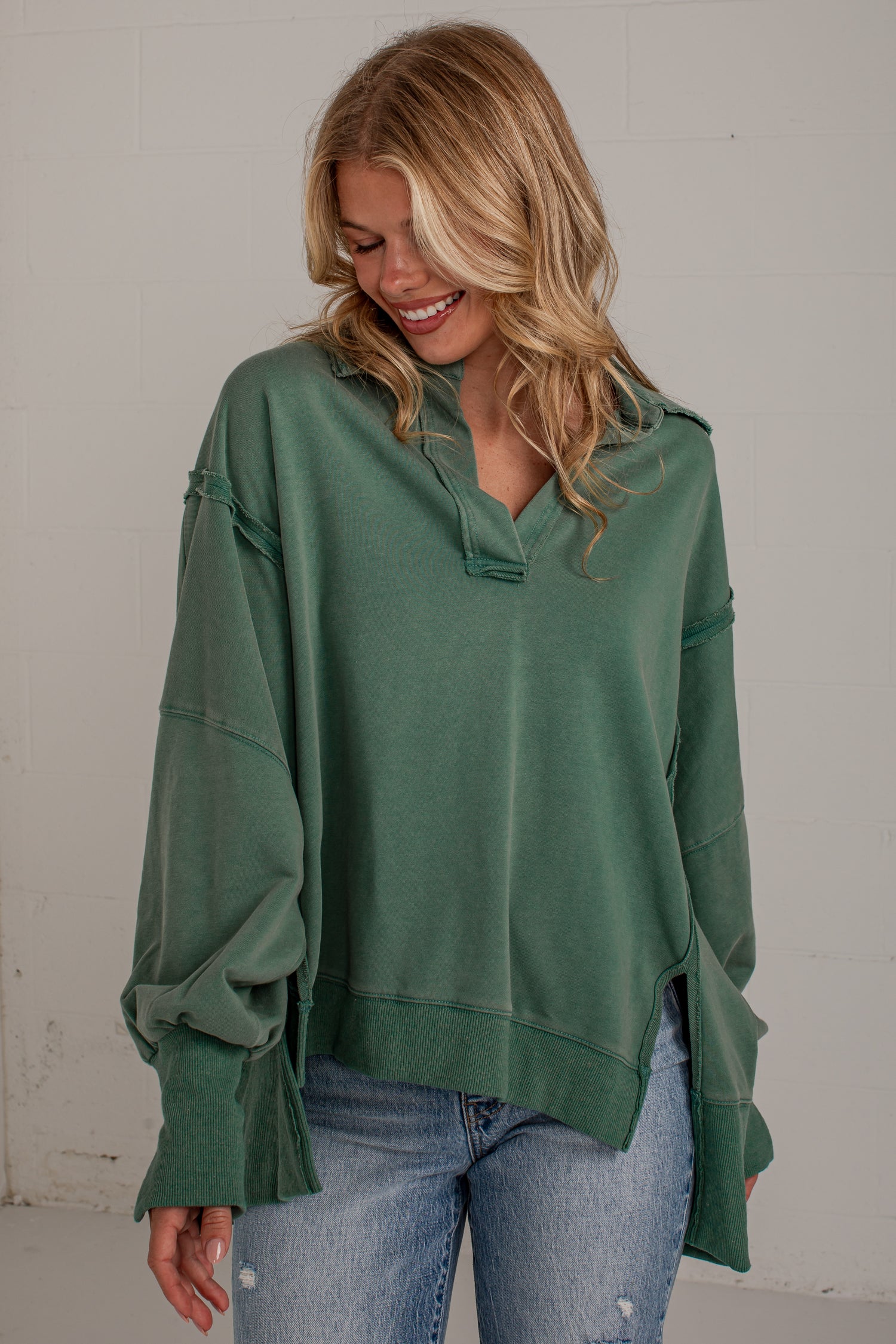 A woman wearing an oversized sage green knit top with unique ruffled cuffs and a split neckline, paired with light-wash blue jeans, standing against a light gray wall.