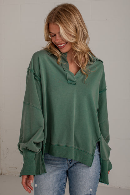 A woman wearing an oversized sage green knit top with unique ruffled cuffs and a split neckline, paired with light-wash blue jeans, standing against a light gray wall.