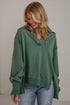 A woman wearing an oversized sage green knit top with unique ruffled cuffs and a split neckline, paired with light-wash blue jeans, standing against a light gray wall.