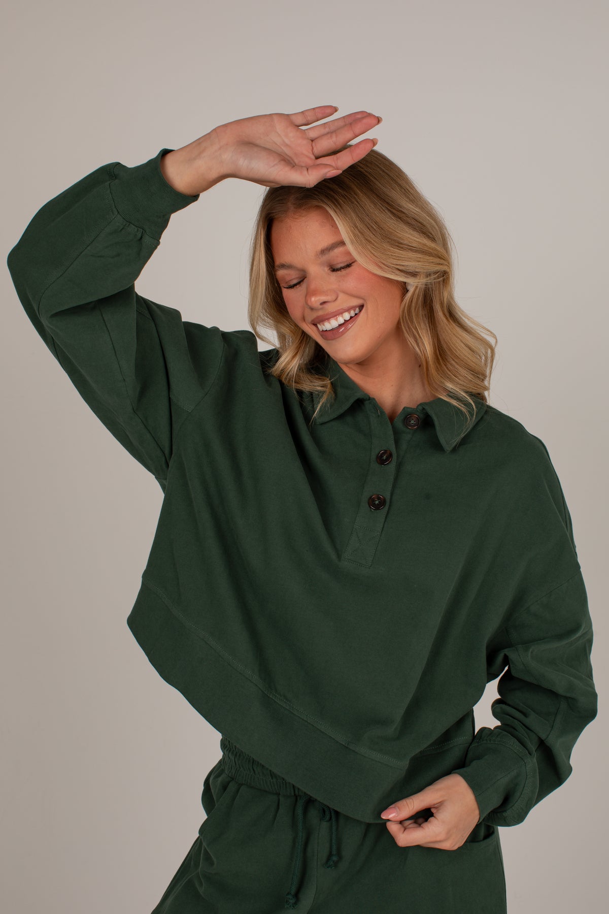 A woman wearing a dark forest green long-sleeve collared pullover top with a button placket, posing with one hand on her head against a solid light gray background.