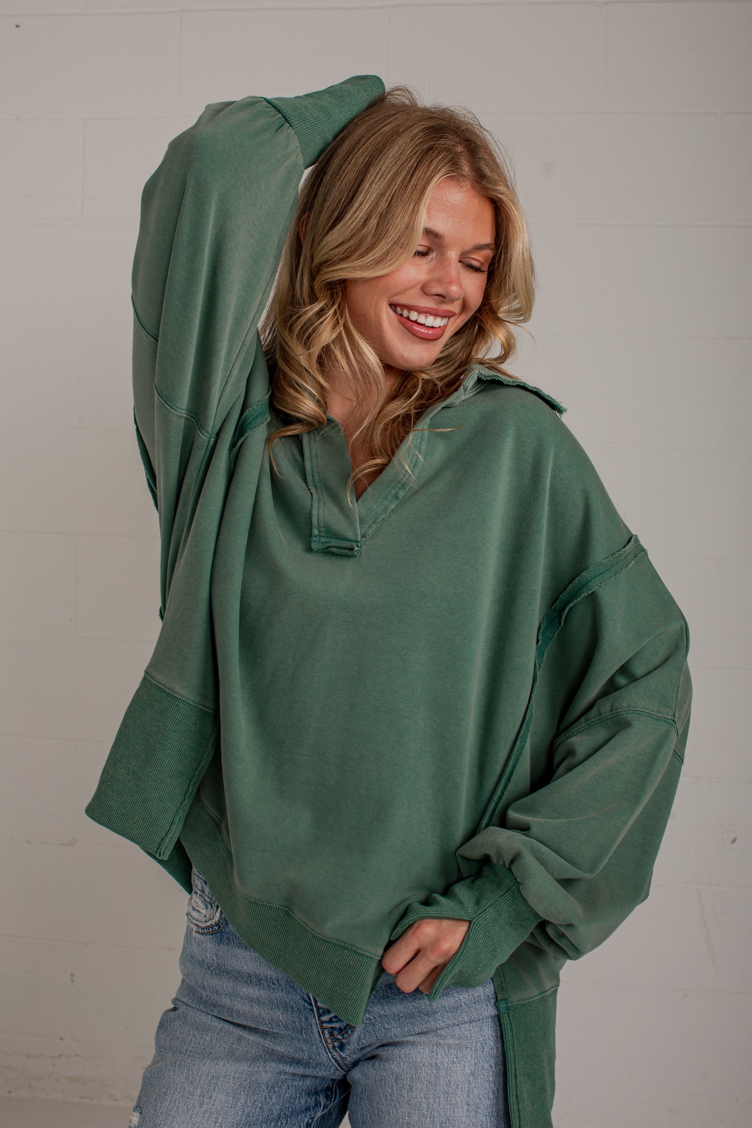 A woman wearing an oversized sage green pullover sweatshirt with a half-placket and drawstring, paired with light-wash blue jeans, posing against a light gray brick wall.