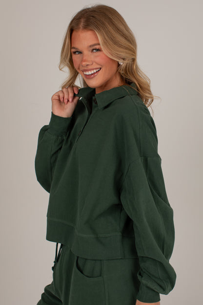 A smiling blonde woman wearing a dark forest green cropped hoodie and matching sweatpants, posing against a solid light gray studio background.