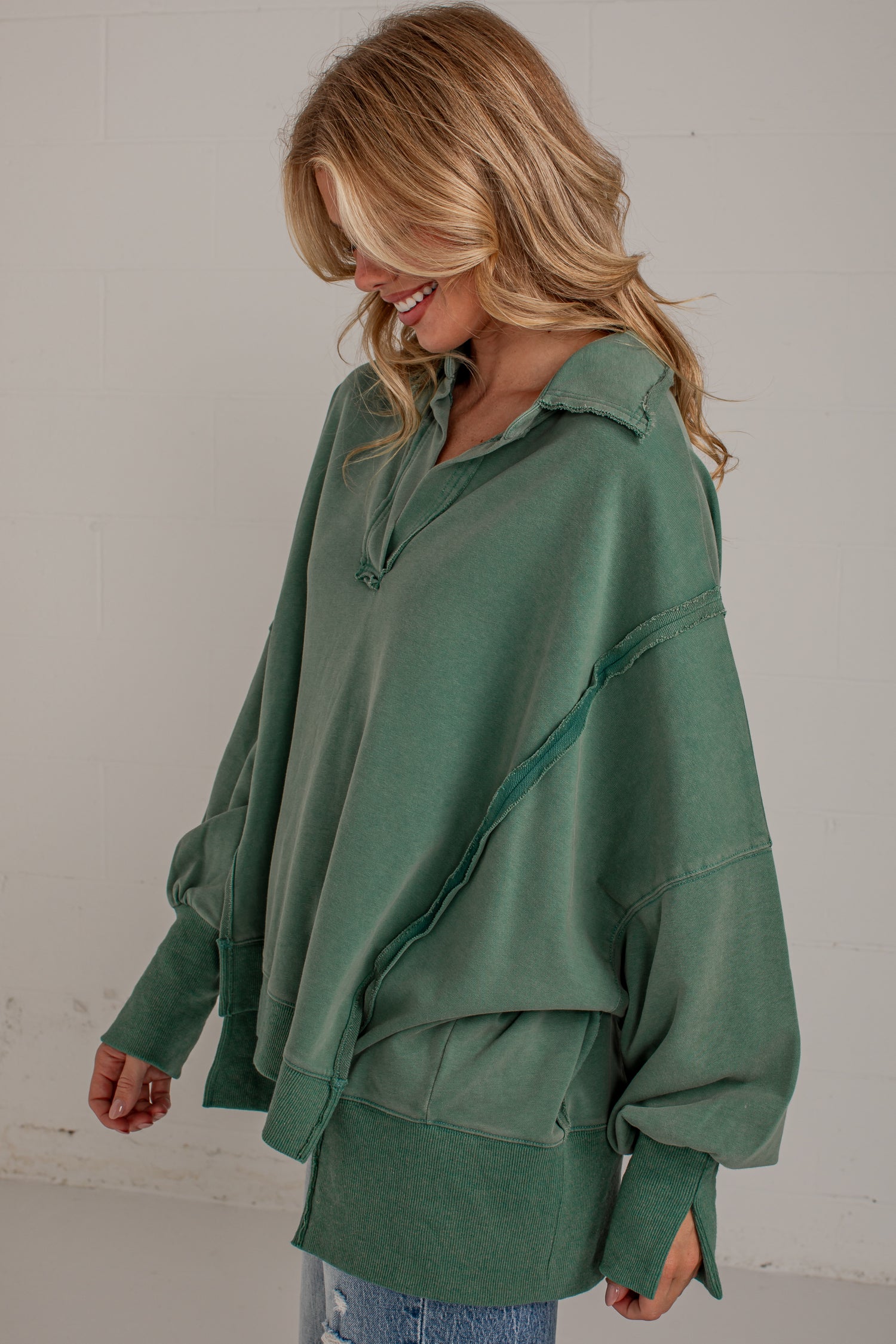 A woman wearing an oversized sage green long-sleeve top with diagonal seam details, shown from the waist up against a light gray wall.
