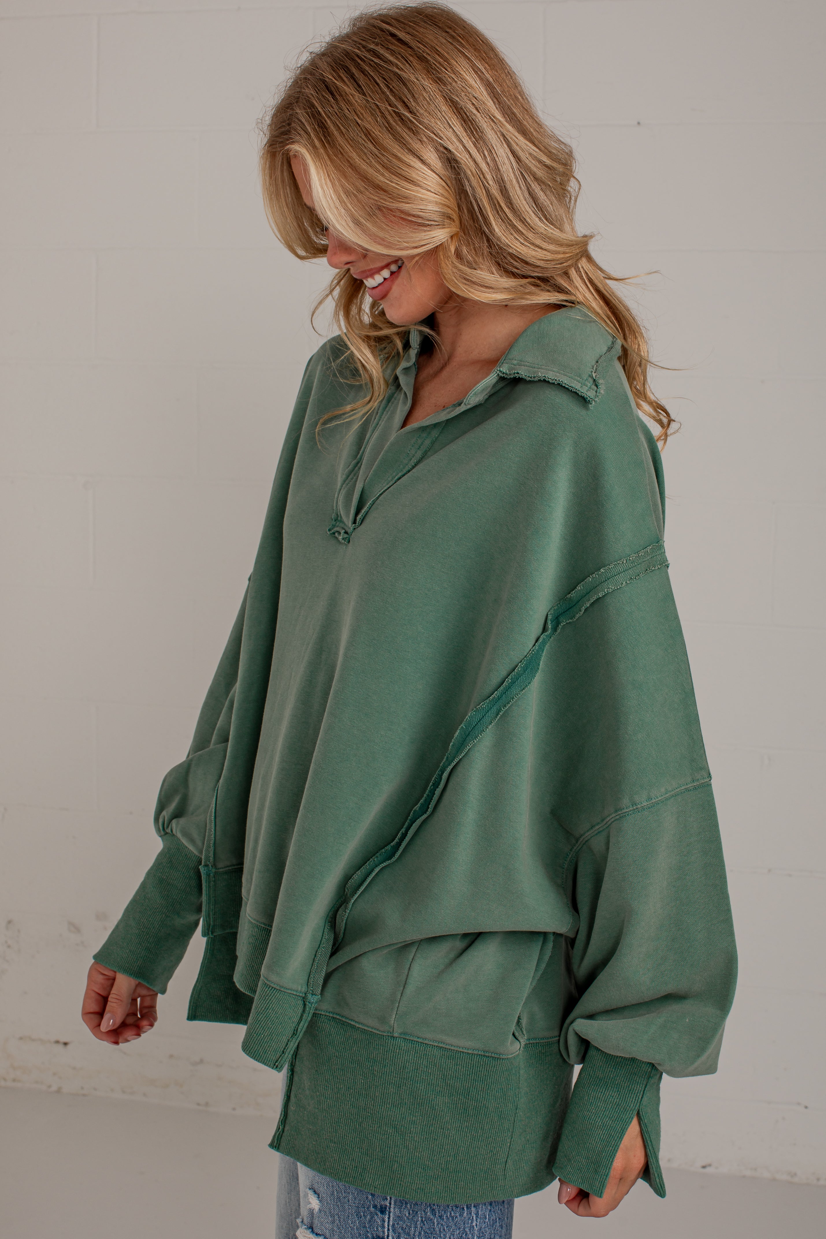 A woman wearing an oversized sage green long-sleeve top with diagonal seam details, shown from the waist up against a light gray wall.