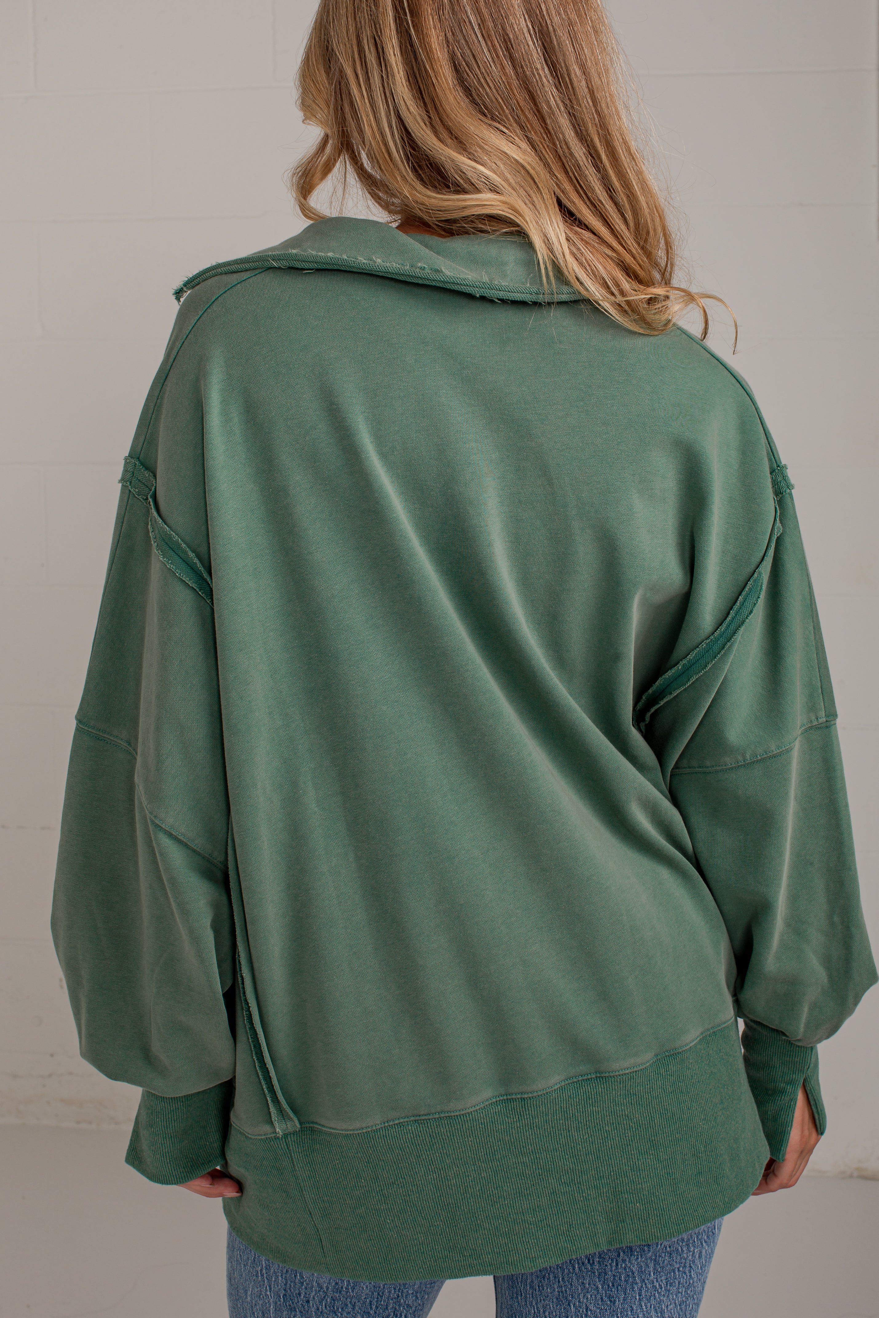 A woman viewed from behind wearing an oversized sage green sweatshirt with unique diagonal seam details and a wide collar, paired with blue jeans.