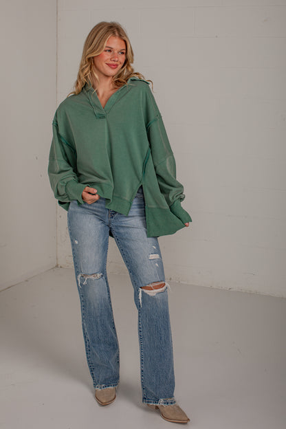 A woman modeling an oversized sage green long-sleeve sweatshirt with a split neckline, paired with distressed light blue jeans and beige ankle boots, standing against a white studio wall.
