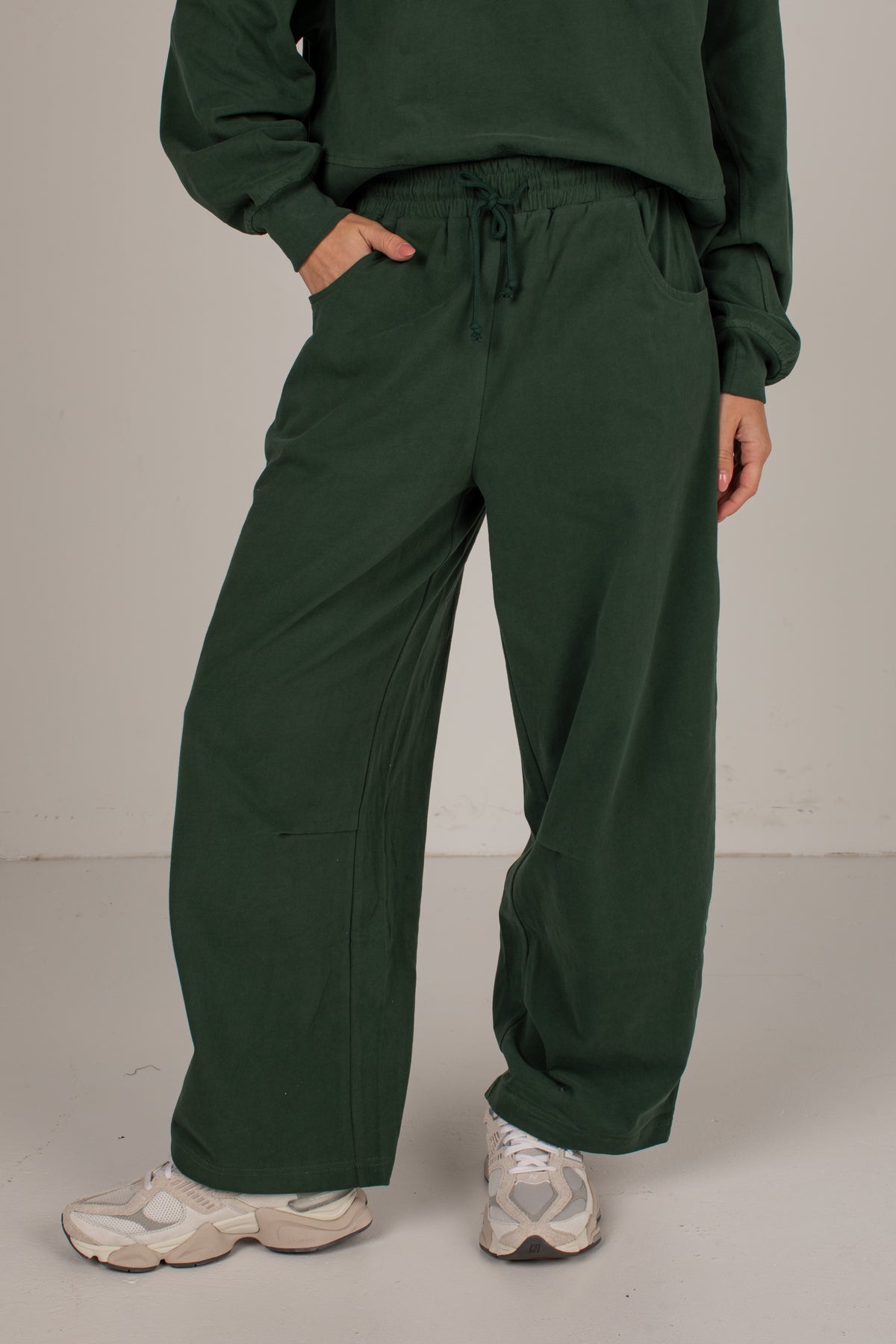 A person wearing dark forest green wide-leg sweatpants with a drawstring waist and side pockets, paired with a matching green sweatshirt and beige sneakers, standing against a neutral studio backdrop.