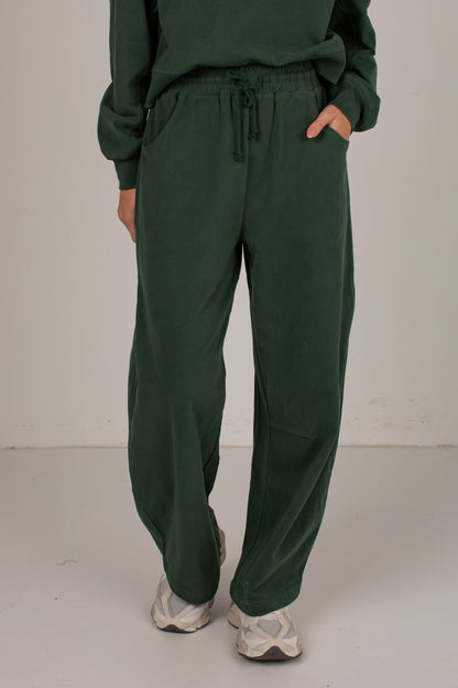 A person wearing dark green drawstring sweatpants with a matching sweatshirt, standing against a plain light gray studio background.