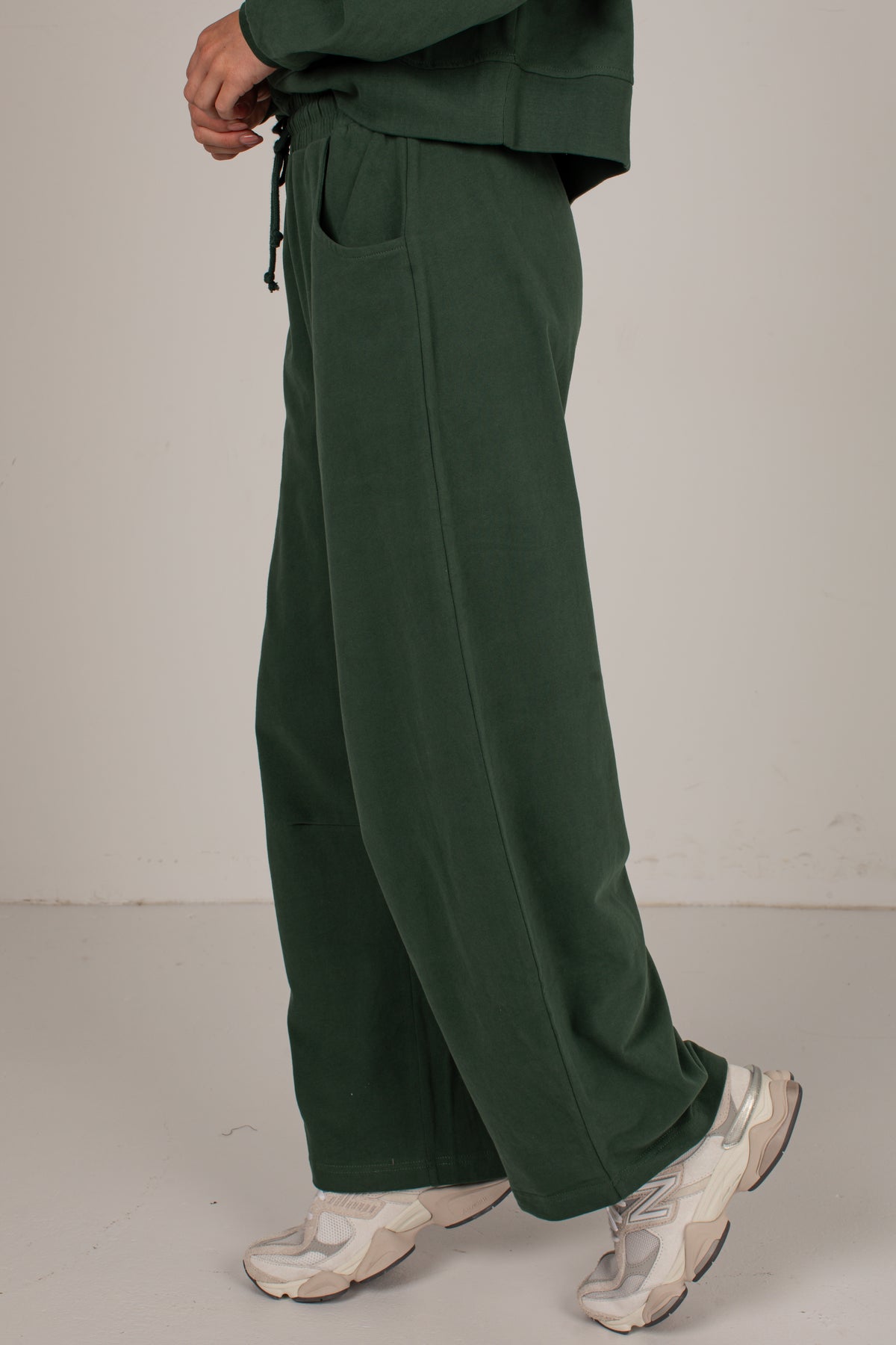 A person wearing dark forest green wide-leg sweatpants with a drawstring waist and side pockets, paired with beige and white sneakers, shown from the waist down against a plain light gray studio background.