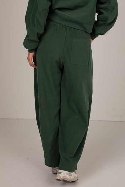 A person shown from the back wearing dark green wide-leg pants with a back pocket, a matching dark green sweatshirt, and light-colored sneakers, standing against a plain off-white studio background.