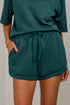 A person wearing dark teal green drawstring shorts with side pockets and a matching top, shown from the chest down against a light neutral wall.