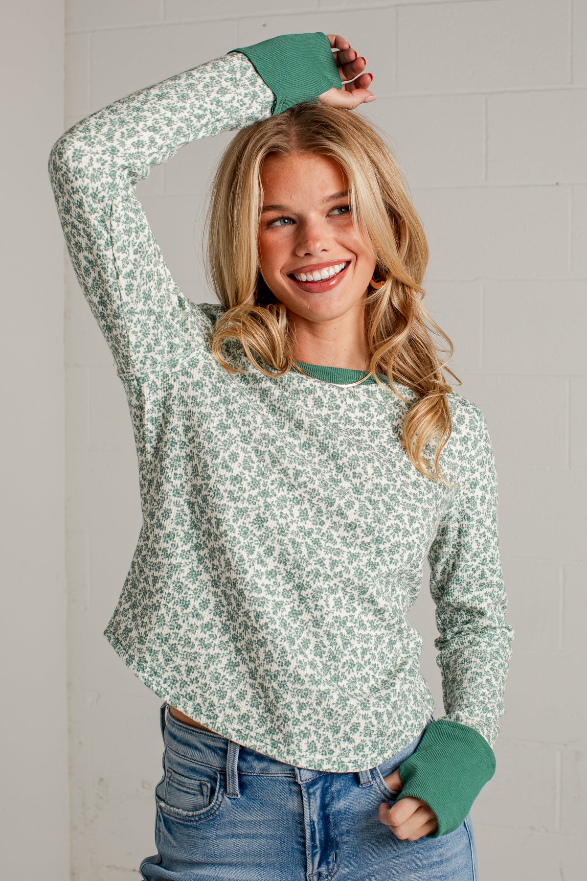 A smiling blonde woman wearing a sage green and white floral patterned long-sleeve shirt with solid green ribbed cuffs and collar, paired with blue jeans, posing against a light gray brick wall.
