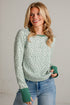 A woman wearing a sage green and white floral patterned long-sleeve sweater with solid green ribbed cuffs and collar, paired with blue jeans, standing against a light gray brick wall.