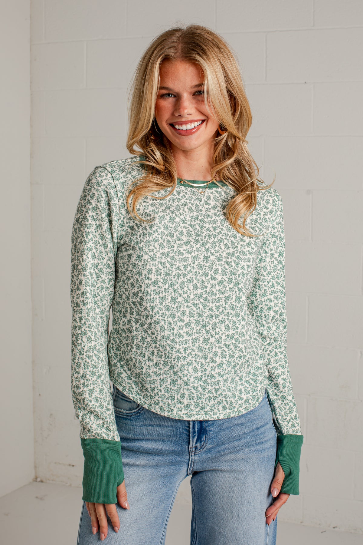 A smiling blonde woman wearing a sage green and white floral patterned long-sleeve top with solid green ribbed cuffs, paired with light blue jeans, standing against a white brick wall.