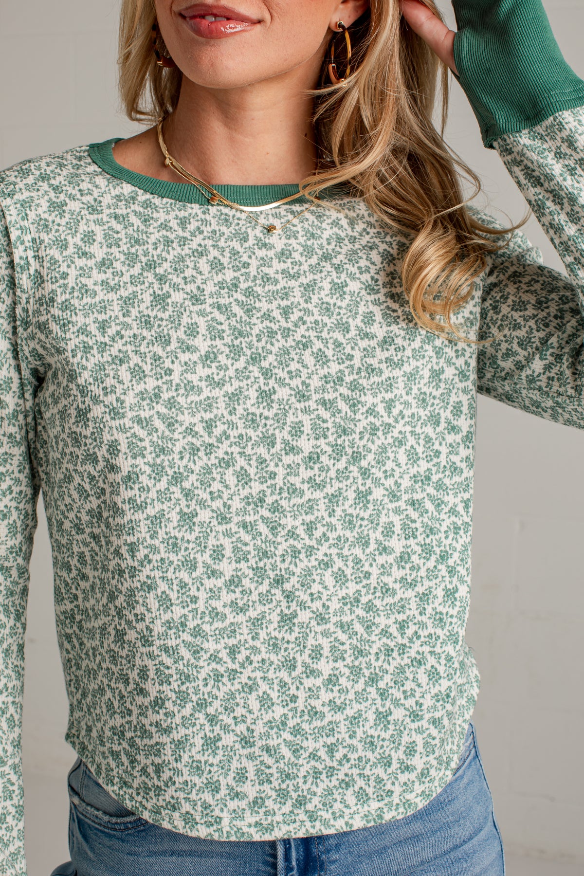 A woman wearing a long-sleeve crewneck top with a green and white floral pattern, paired with blue jeans and gold jewelry, shown in a close-up shot.