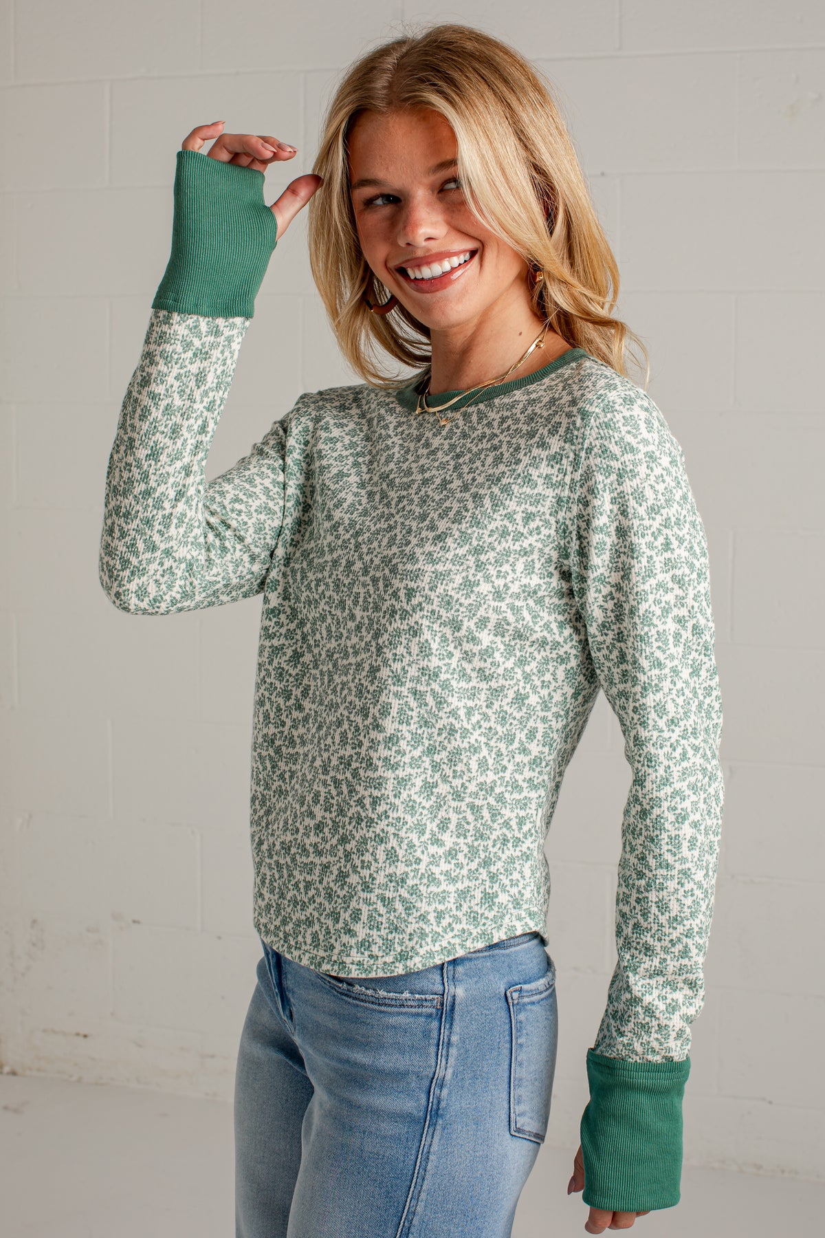 A woman wearing a sage green and white floral patterned long-sleeve top with solid green ribbed cuffs, paired with light blue jeans, standing against a white brick wall.