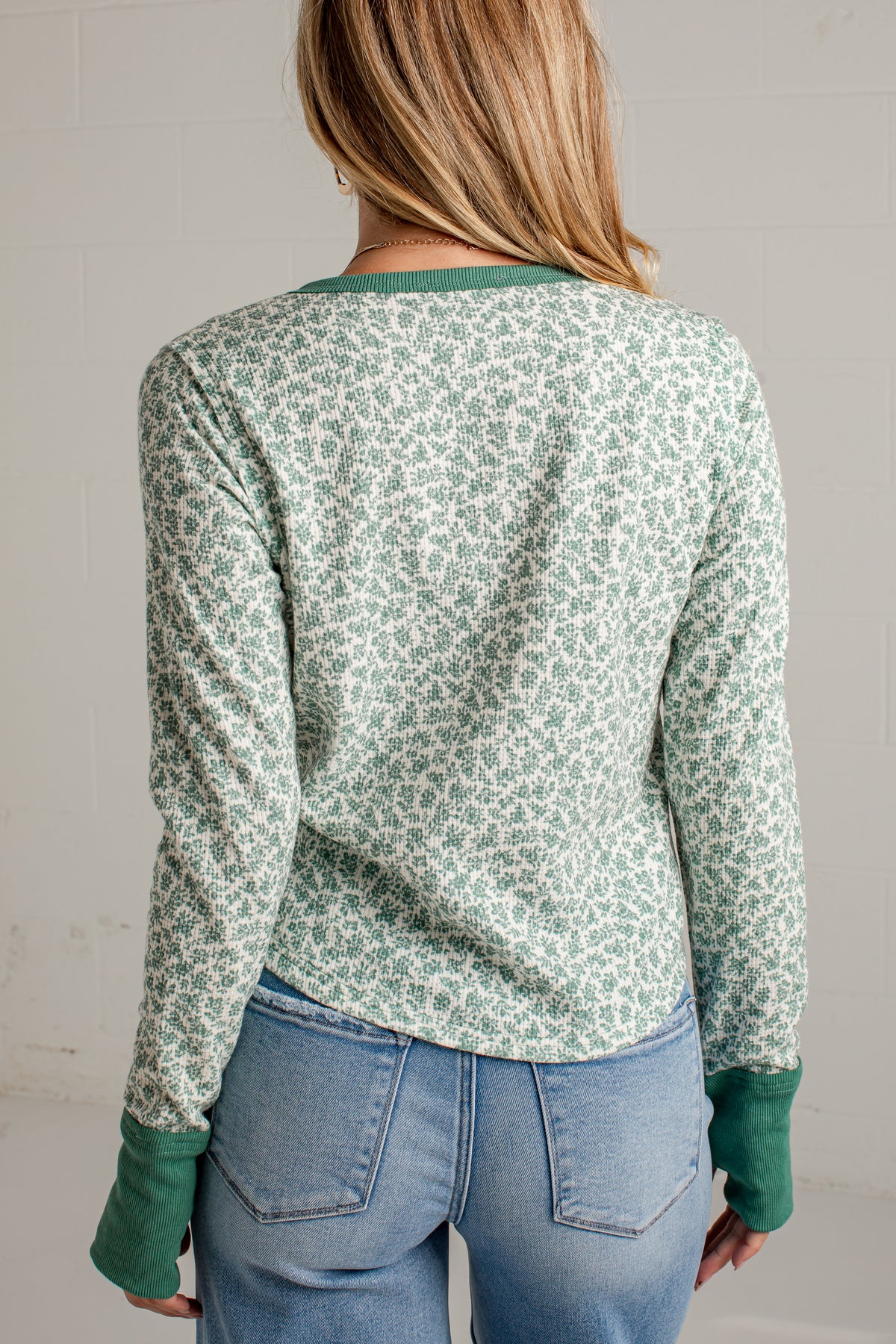 A woman viewed from the back wearing a long-sleeve knit top with a light sage green and white floral pattern and solid green ribbed collar, cuffs, and hem, paired with light blue jeans.