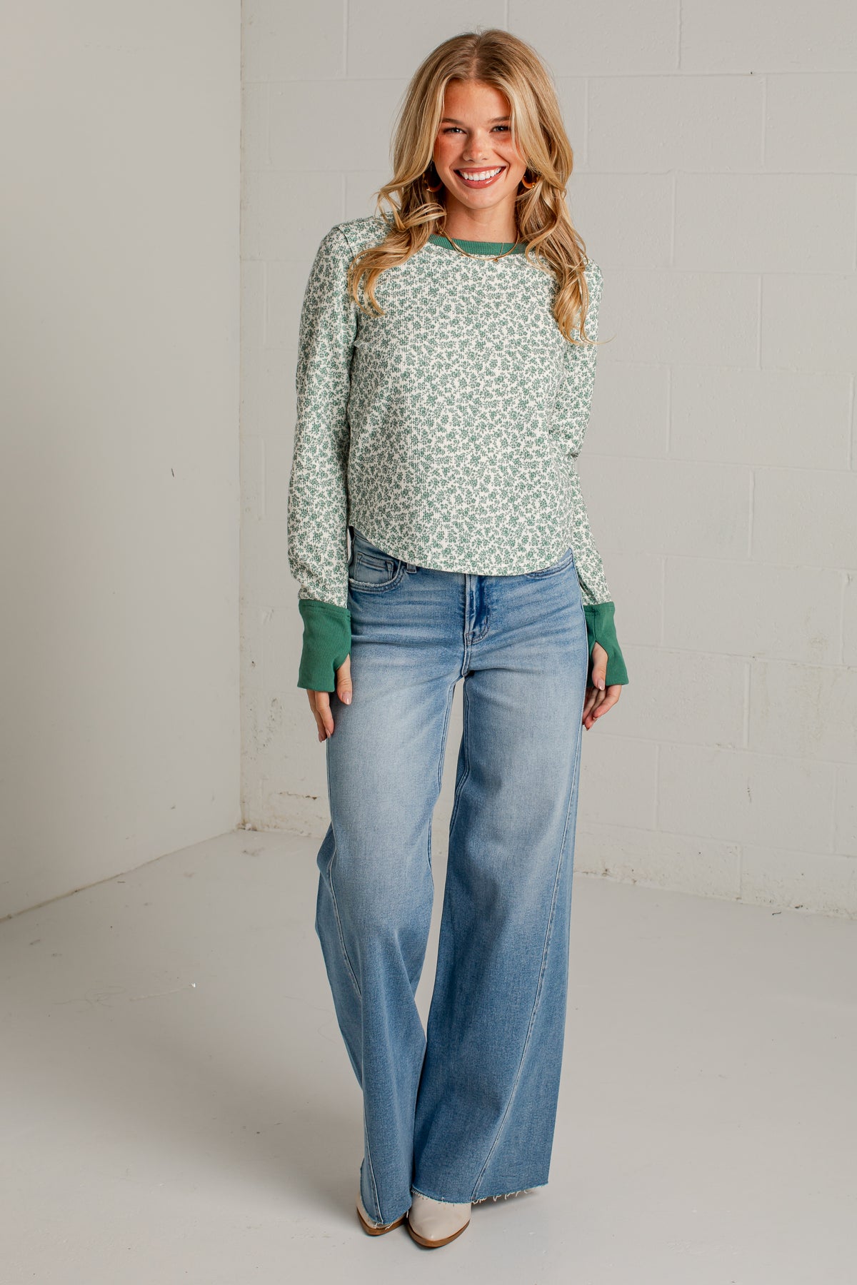 A woman modeling a light green and white patterned long-sleeve top with contrasting green cuffs, paired with wide-leg blue jeans, standing in a bright studio with white walls.