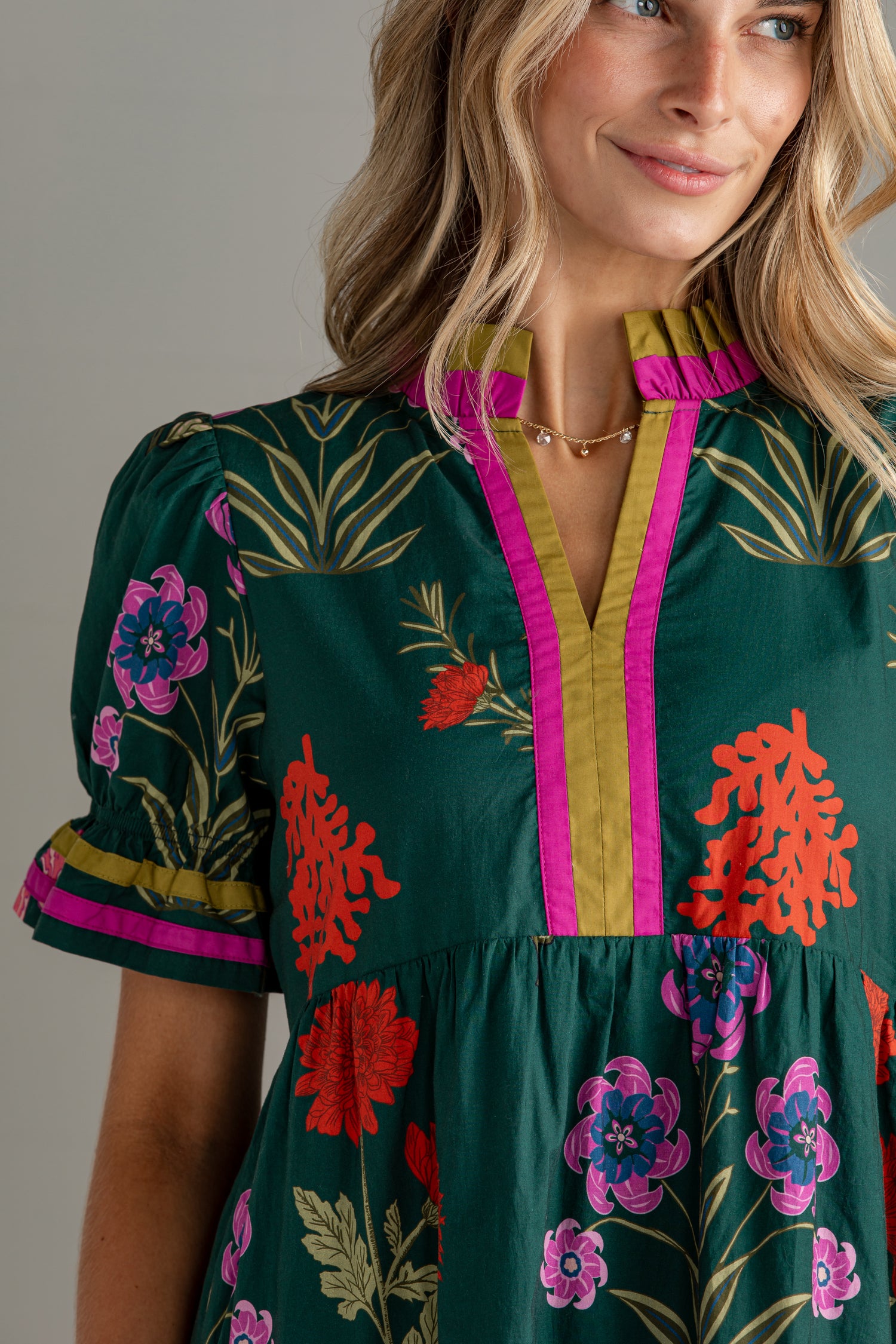 A close-up of a woman wearing a dark green short-sleeve dress with a colorful floral print, featuring a yellow and pink ruffled V-neckline.