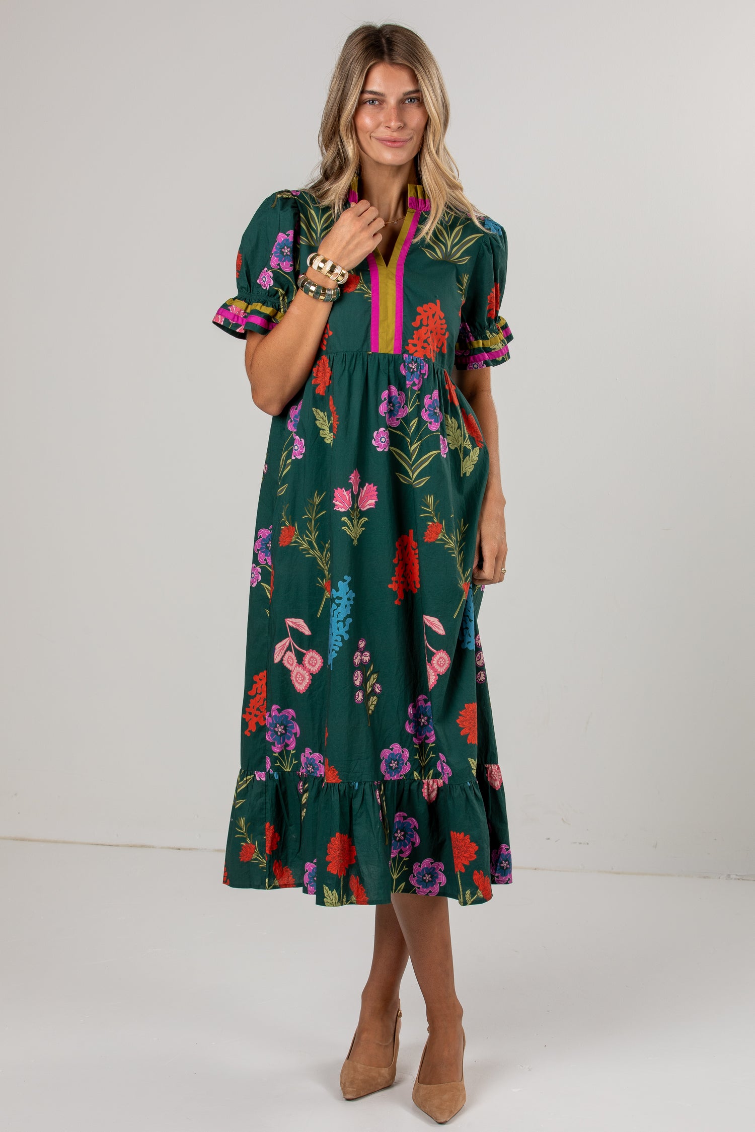 A woman modeling a dark green midi dress with a colorful floral print, puff sleeves, and a pink and yellow striped V-neckline, set against a plain light gray studio background.