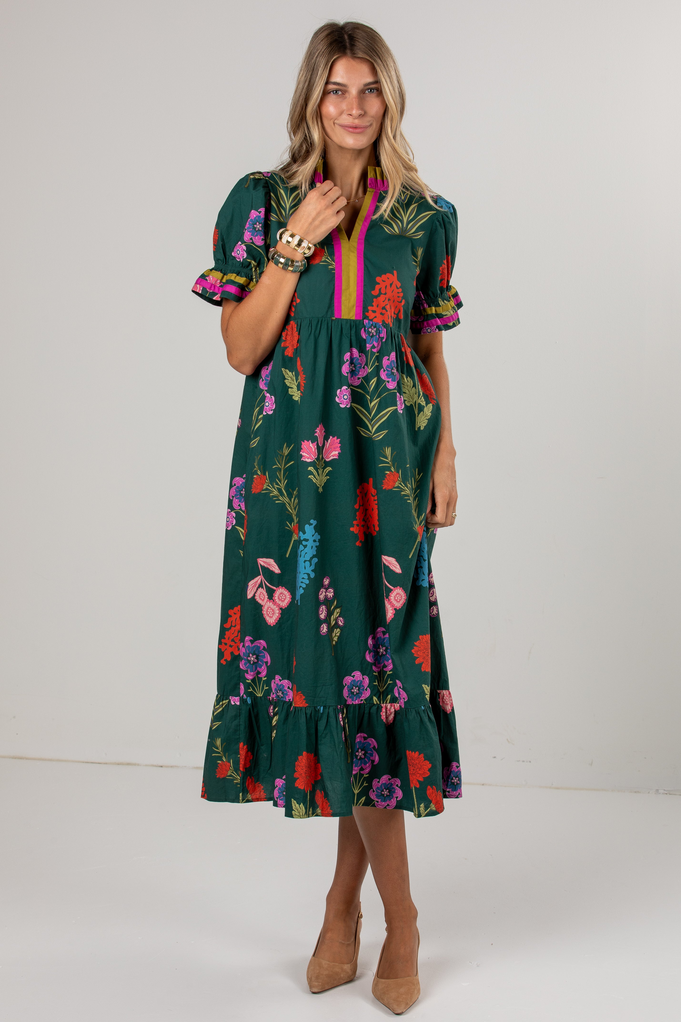 A woman modeling a dark green midi dress with a colorful floral print, puff sleeves, and a pink and yellow striped V-neckline, set against a plain light gray studio background.