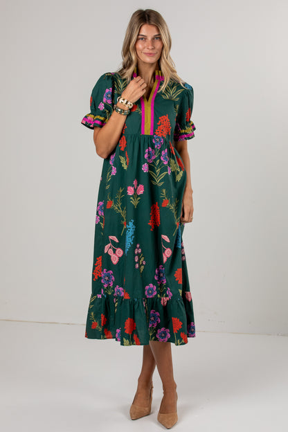 A woman modeling a dark green midi dress with a colorful floral print, puff sleeves, and a pink and yellow striped V-neckline, set against a plain light gray studio background.