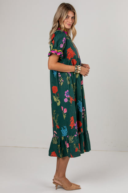 A woman modeling a dark green midi dress with a colorful floral print, featuring short puff sleeves with ruffled trim and a tiered ruffle hem, paired with tan heels against a plain light gray studio background.