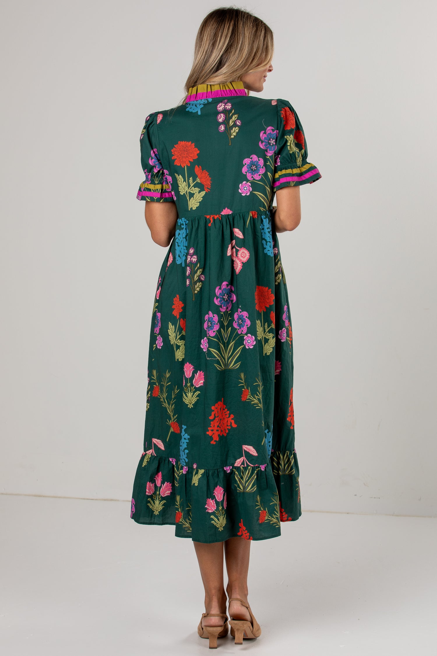 A woman viewed from the back wearing a dark green midi dress with a colorful floral print, short puff sleeves, and a ruffled hem, paired with tan heeled sandals against a plain light gray studio background.