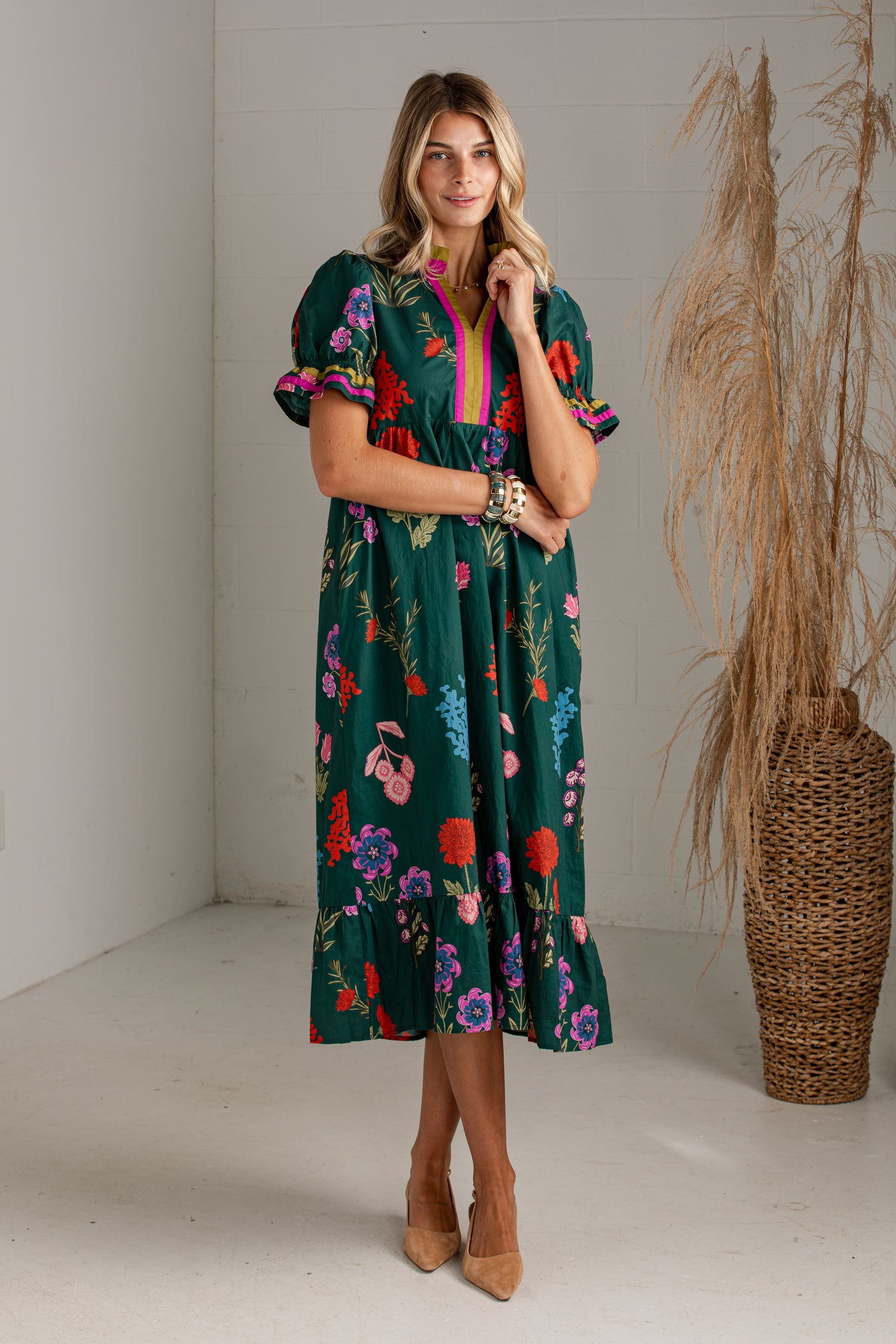 A woman models a dark green midi dress with a colorful floral print, featuring puff sleeves and a bright pink V-neck trim, standing in a studio with a pampas grass vase in the background.
