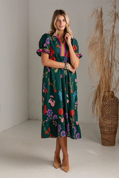 A woman models a dark green midi dress with a colorful floral print, featuring puff sleeves and a bright pink V-neck trim, standing in a studio with a pampas grass vase in the background.