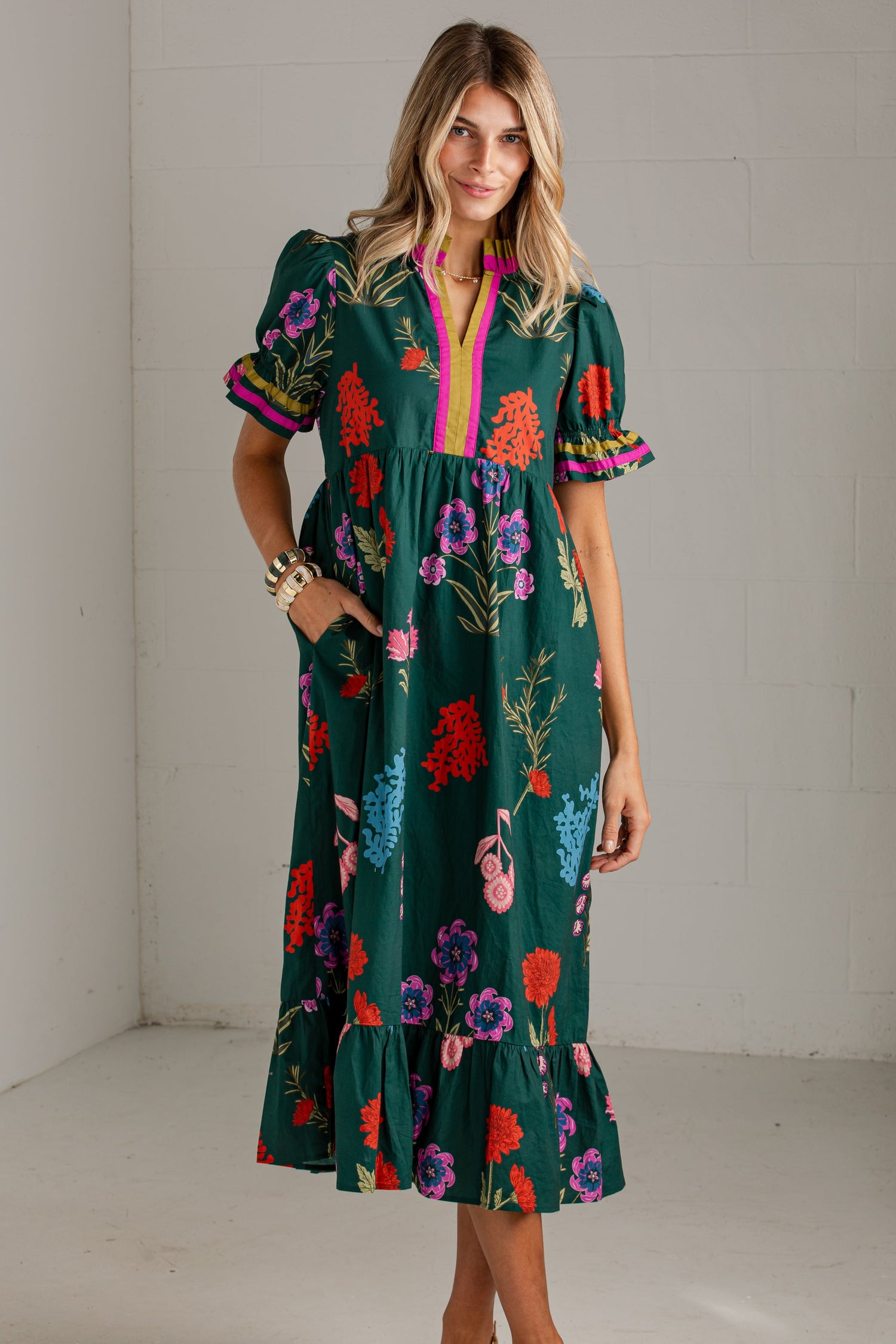 A woman modeling a dark green midi dress with a vibrant multicolor floral print, short puff sleeves, and a pink trim along the split neckline, standing in a white studio corner.