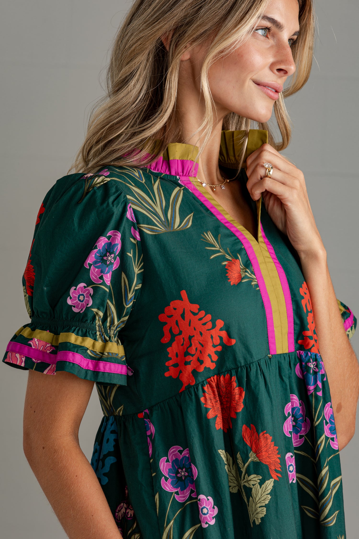 A woman wearing a dark green floral dress with vibrant pink, red, and purple flowers, featuring a yellow and pink ruffled collar and puff sleeves.