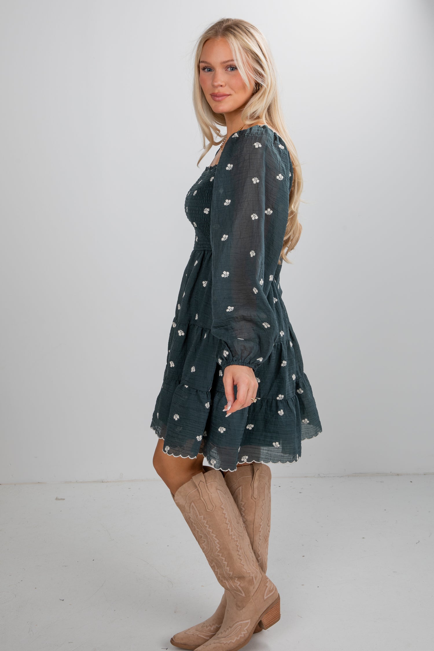 Woman wearing a dark green dress with white patterns and beige knee-high boots on a plain background