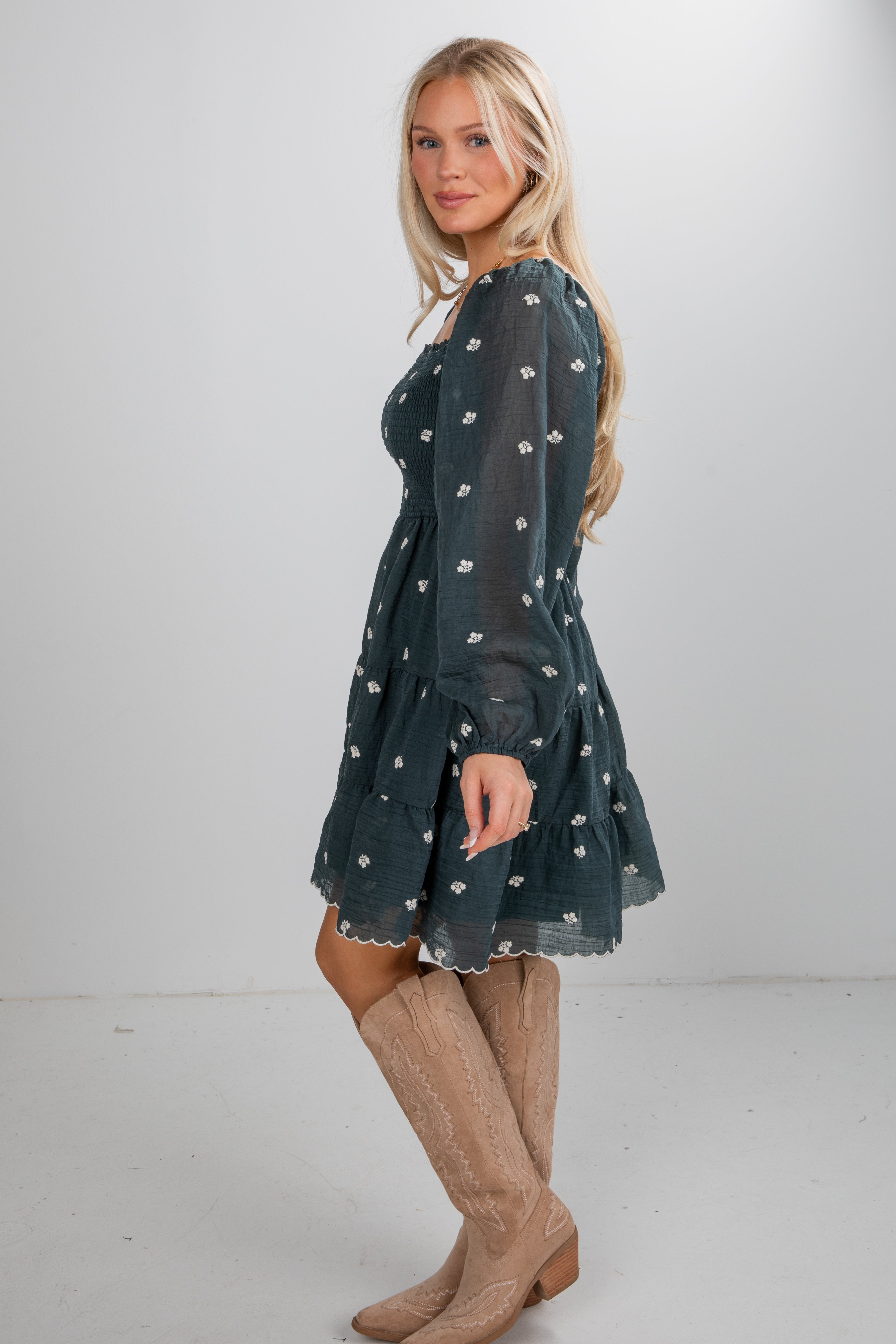 Woman wearing a dark green dress with white patterns and beige knee-high boots on a plain background