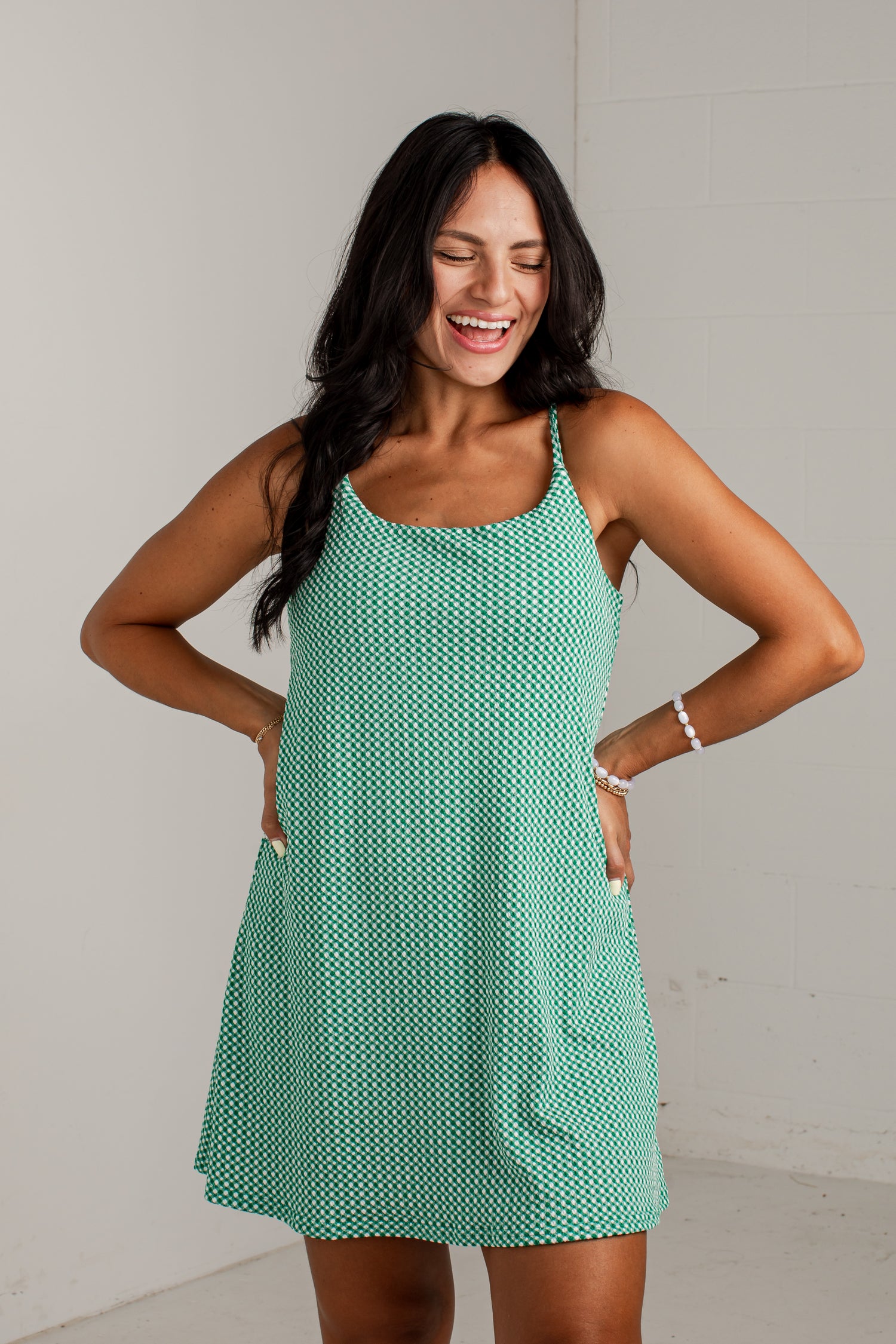 A woman modeling a sleeveless, scoop-neck mini dress with thin straps and side pockets, featuring a mint green and white geometric pattern, set against a plain light gray wall.