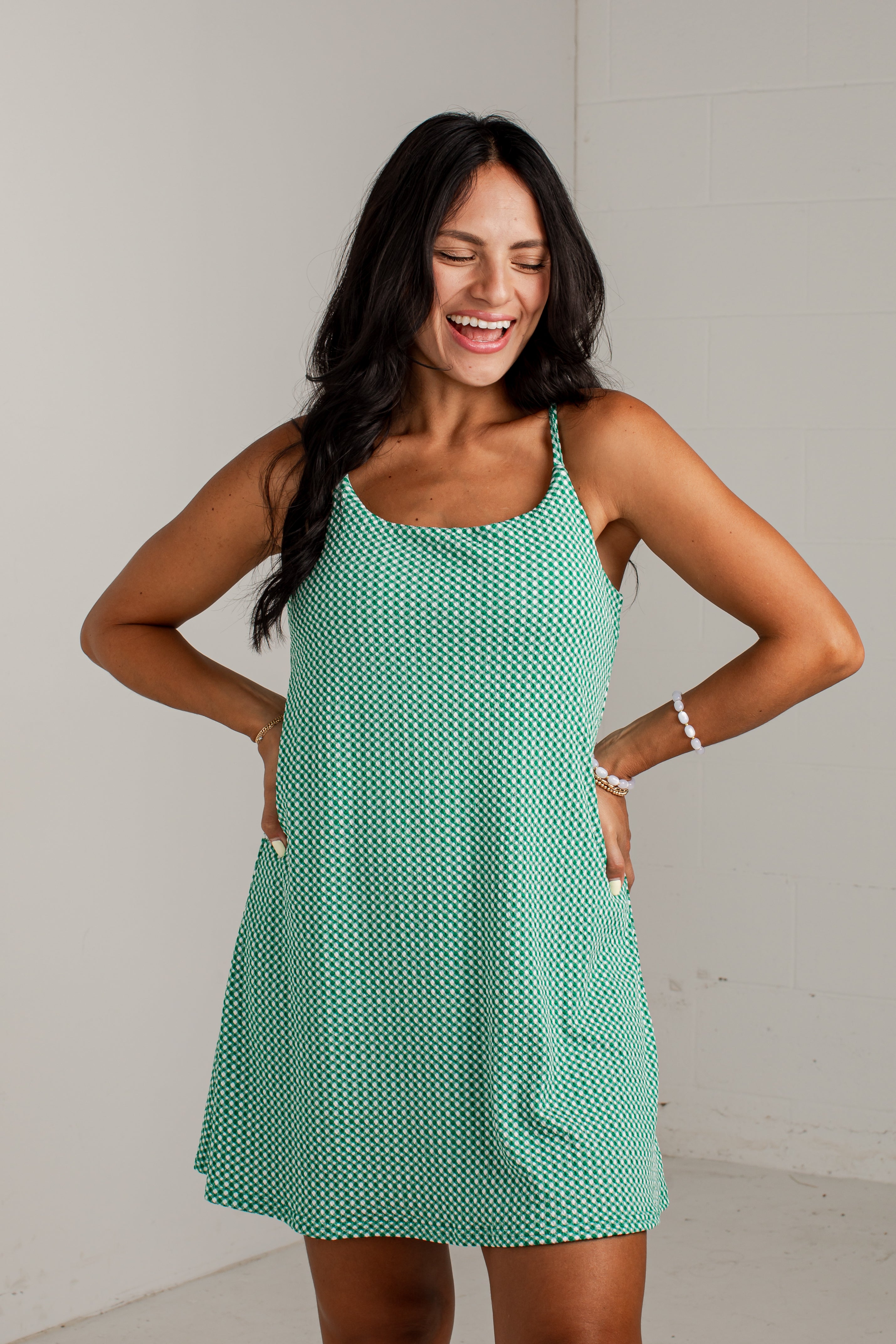 A woman modeling a sleeveless, scoop-neck mini dress with thin straps and side pockets, featuring a mint green and white geometric pattern, set against a plain light gray wall.