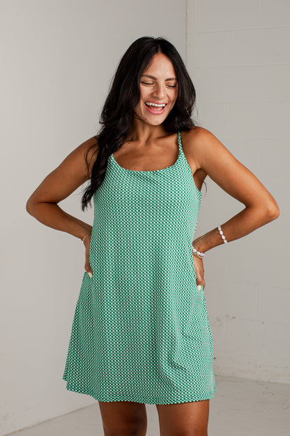 A woman modeling a sleeveless, scoop-neck mini dress with thin straps and side pockets, featuring a mint green and white geometric pattern, set against a plain light gray wall.