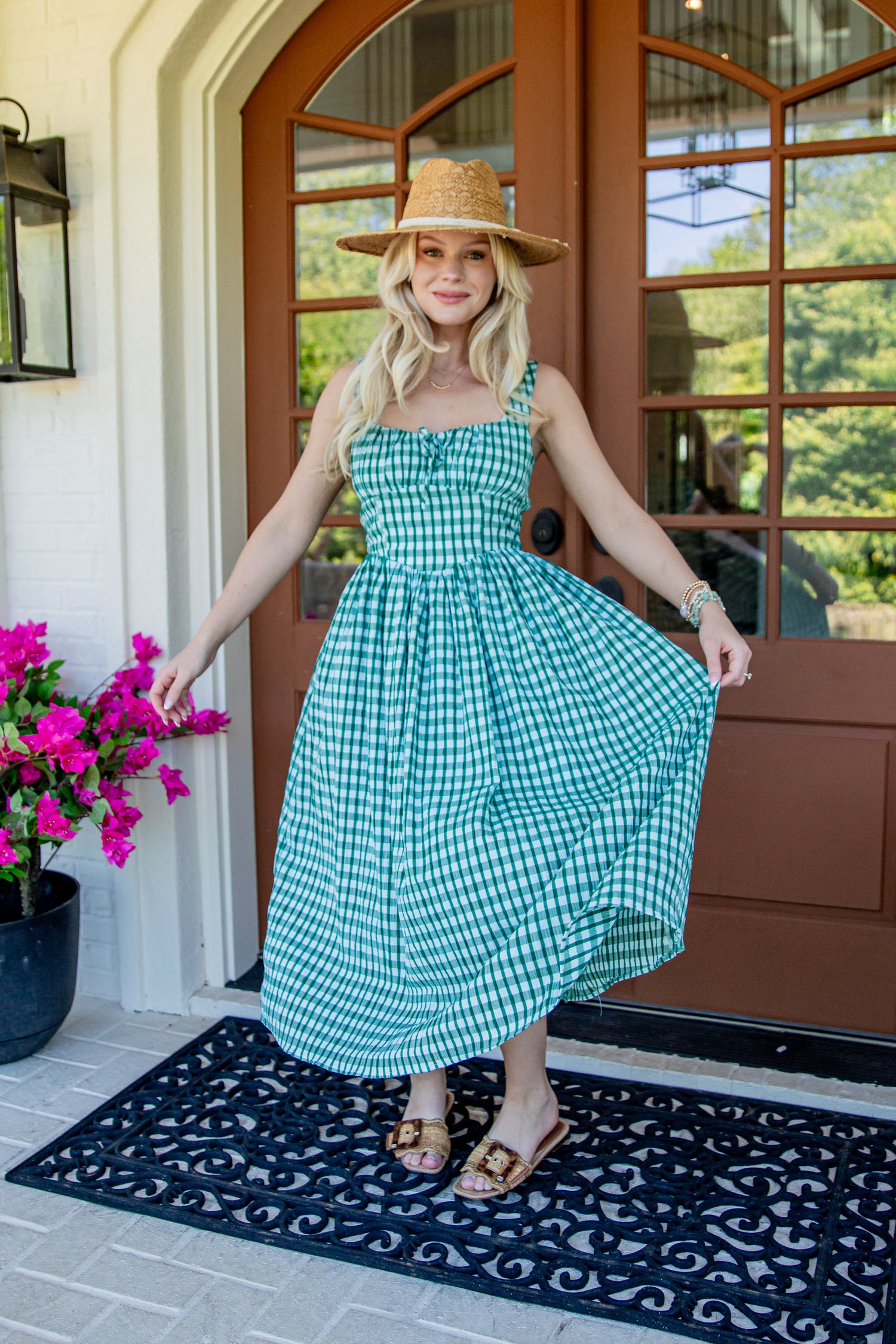 A woman wearing a green and white gingham midi dress with a straw hat stands on a front porch holding out her dress to show the flow. The porch has a brown wooden door, a black patterned doormat, and a potted plant with bright pink flowers.