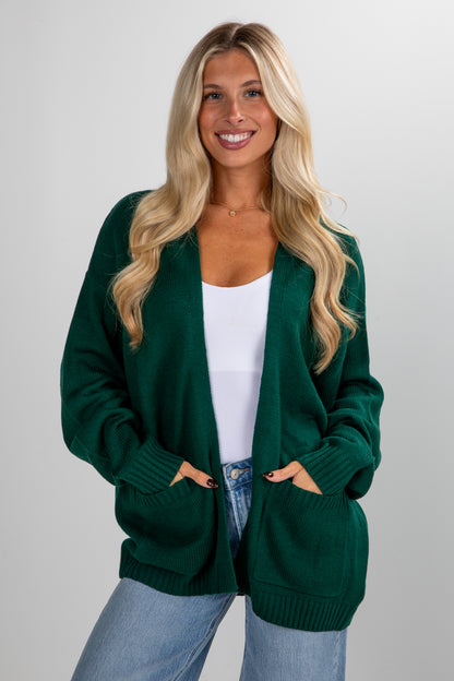 Woman wearing a green cardigan over a white top and blue jeans on a plain background
