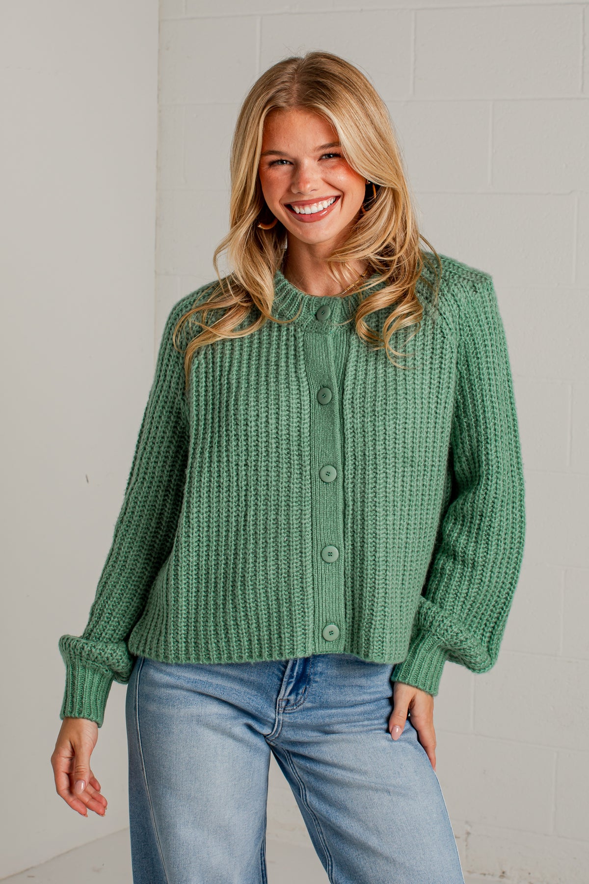 A smiling blonde woman wearing a sage green ribbed knit cardigan with button-front closure and light blue jeans, standing against a light gray brick wall.