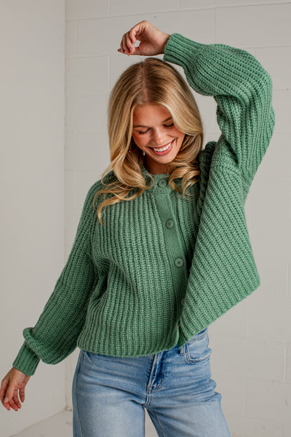 A woman wearing a green chunky knit cardigan sweater and light blue jeans, posing in front of a white brick wall.