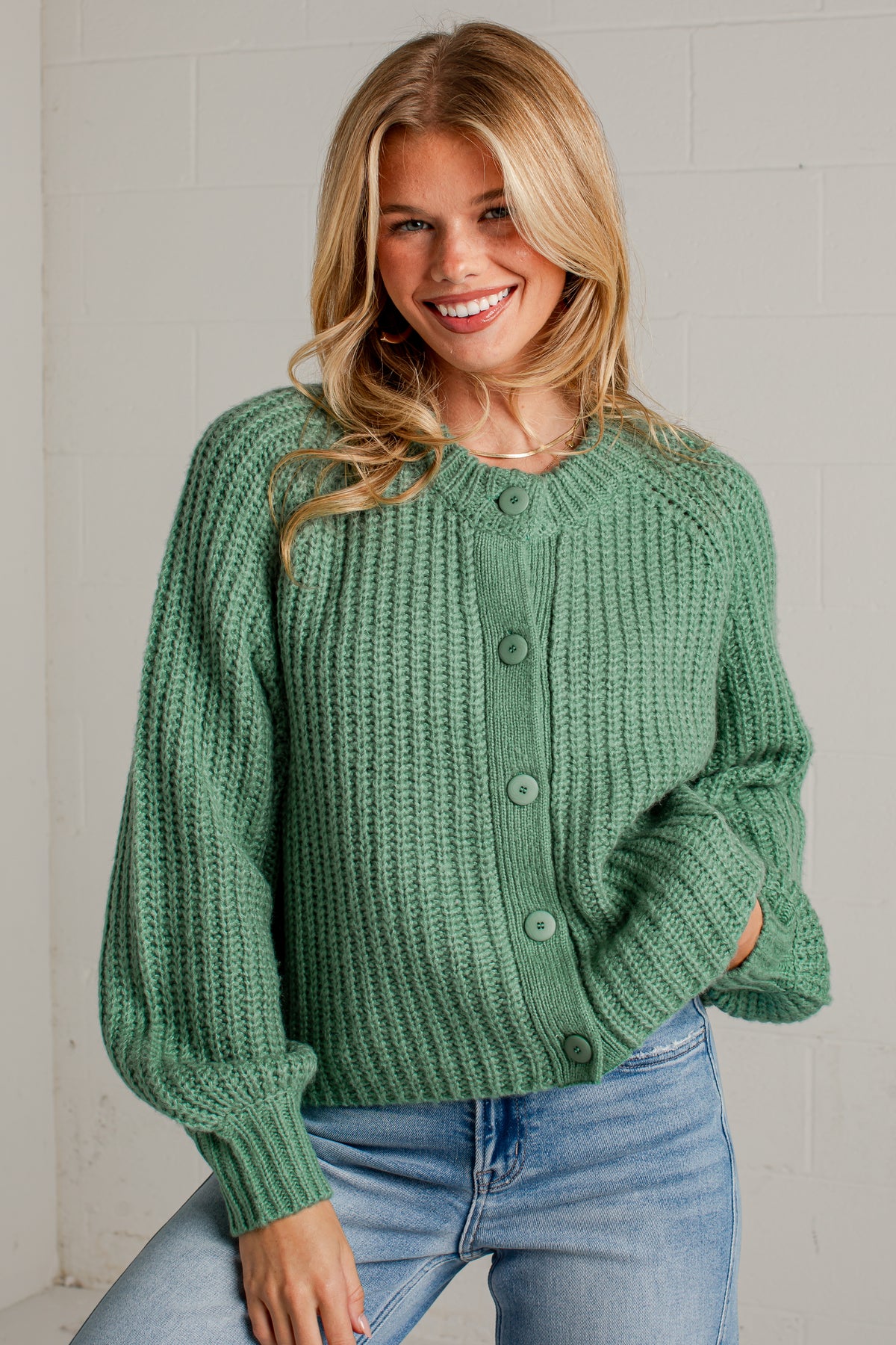 A smiling blonde woman wearing a chunky knit sage green cardigan sweater with button-front closure and light blue jeans, posed against a light gray brick wall.
