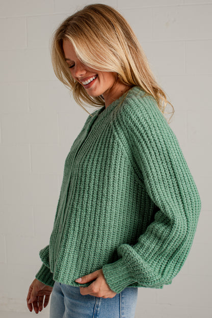 A woman wearing a chunky knit sage green sweater and light blue jeans, smiling and looking down against a white brick wall background.