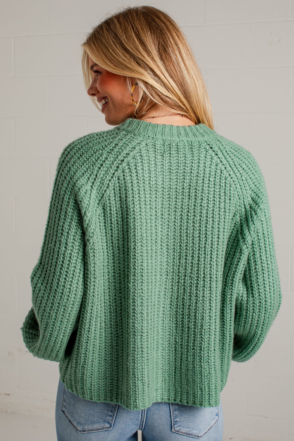 A woman wearing a sage green chunky knit sweater, shown from the back against a white brick wall.